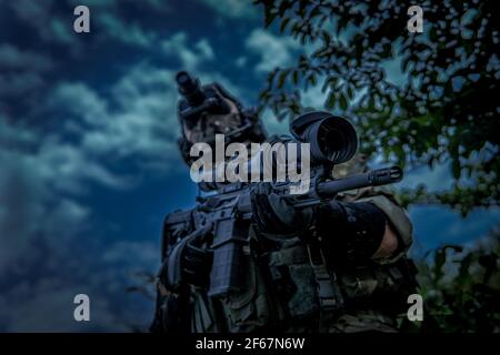 Il soldato sta tenendo un'arma con una vista telescopica. Vista dall'angolo inferiore. Dispositivo di visione notturna al buio. Foto Stock