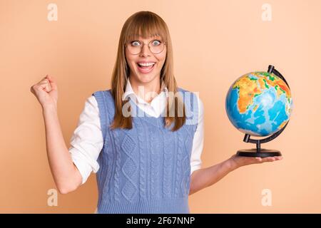 Ritratto fotografico di ragazza stupita che tiene il globo a scuola gesturing come vincitore in bicchieri isolati su sfondo di colore beige pastello Foto Stock