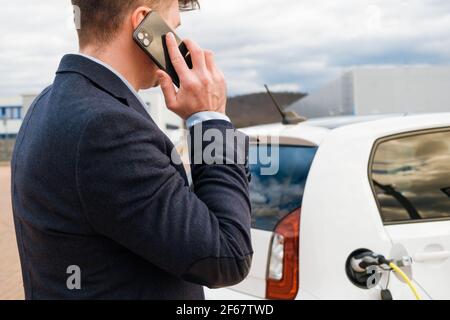 Uomo d'affari in piedi vicino all'elettro di carica e che parla sopra lo smartphone Foto Stock