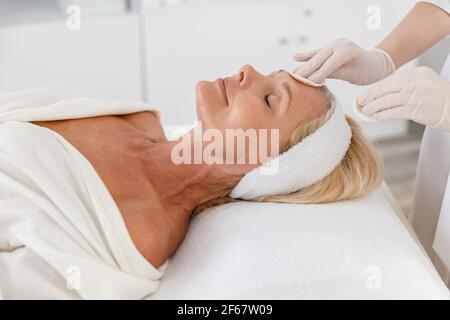 Primo piano trattamento di bellezza viso spa, cura della pelle. Donna anziana che si cura del viso da estetista al salone termale, vista laterale Foto Stock