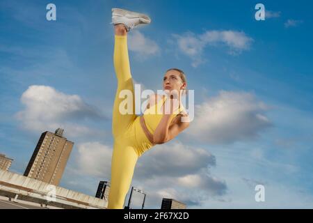 Attraente donna sportiva che indossa activewear giallo che pratica l'esercizio di alto calcio all'esterno in un ambiente urbano. Foto Stock