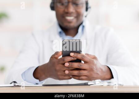 Consultazione online. Medico di sesso maschile afroamericano che usa lo smartphone sul posto di lavoro, messaging con il suo paziente Foto Stock