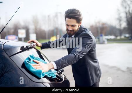 Elegante giovane uomo in tuta che pulisce il finestrino posteriore della vettura con un panno in microfibra blu. Foto Stock