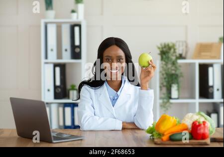 Donna afroamericana dietista tenendo mela e sorridendo alla macchina fotografica in un ufficio moderno Foto Stock