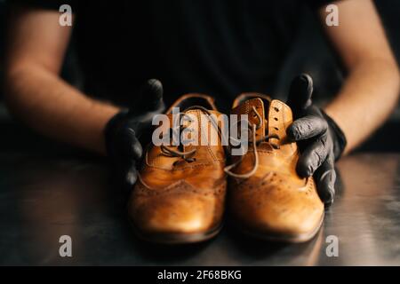 Vista frontale ravvicinata delle mani del calzolaio nero guanti che tengono vecchie scarpe in pelle marrone chiaro Foto Stock