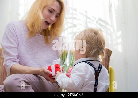 Il ragazzino si congratula con la madre per la vacanza, le dà fiori e un regalo. Ragazzo in camicia bianca e pantaloni con sospenditori Foto Stock