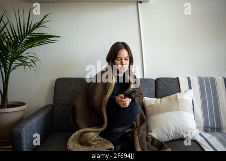 Donna avvolta in coperta seduta sul divano guardando il telefono Foto Stock
