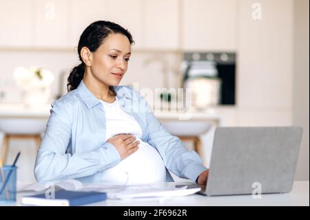Concentrato bella corsa mista riuscita donna incinta, freelancer, manager o designer che lavorano a distanza al laptop, controllare il rapporto di affari o il progetto di progettazione, stroking ventre, sorridente Foto Stock