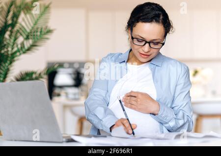 Donna di corsa mista intelligente di successo incinta con occhiali, donna di affari, broker o freelancer, lavorando a distanza, utilizza laptop, prendere appunti e controllare business plan, stracciare il ventre, sorridere Foto Stock