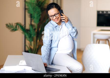 Donna incinta impegnata. Donna di corsa mista incinta di successo con occhiali, manager, broker o freelancer, usa il laptop, parla al telefono con un collega o un cliente, seduto sulla scrivania, sorridente Foto Stock