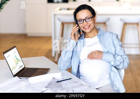 Donna incinta impegnata. Elegante bruna gravidanza sicura e intelligente mista razza donna d'affari femminile, designer o freelance, seduto sul posto di lavoro, parlando sul cellulare con un collega o cliente, sorridendo Foto Stock