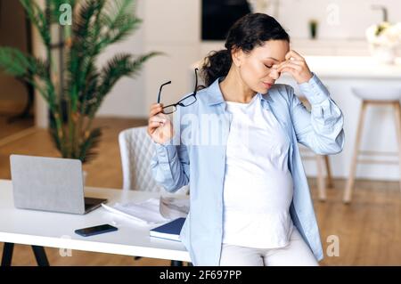 Concetto di overwork. Stressato donna di razza mista incinta, donna d'affari, manager o freelance, lavorando da casa, togliendo gli occhiali, strofinando gli occhi con la mano, in piedi al tavolo sconvolto Foto Stock