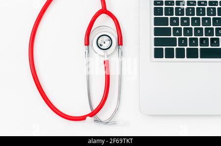 Primo piano di un dettaglio di uno stetoscopio rosso accanto a. un dettaglio di un computer portatile su sfondo bianco Foto Stock