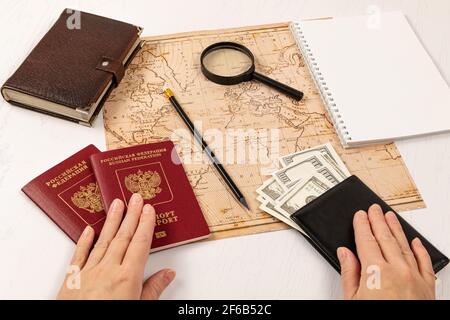 le mani femminili tengono due passaporti e un portafoglio in pelle nera con soldi. su un tavolo di legno bianco si trova una mappa del mondo, una lente d'ingrandimento, una matita, Foto Stock