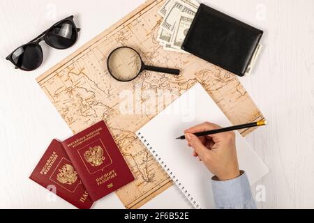 la mano femminile fa note con una matita in un taccuino. su un tavolo di legno bianco si trova una mappa del mondo, due passaporti, una lente d'ingrandimento e una wa nera Foto Stock