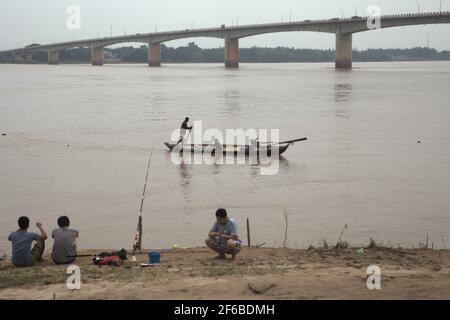 Giovani uomini che pescano dalla riva del fiume come passa una barca da pesca, galleggianti sul fiume Mekong a Kampong Cham, Cambogia. Un'adeguata 'governance delle acque' oltre i confini è fondamentale poiché dighe e sviluppo (ironicamente) minacciano l'ambiente e la vita di milioni di persone, ha scritto Paritta Wangkiat in un'analisi pubblicata da Bangkok Post il 25 marzo 2021. Foto Stock