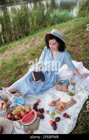 Una donna in cappello di paglia si siede su un picnic dentro un parco sulle rive di un fiume Foto Stock