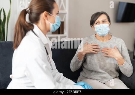 Infermiere in maschera cura di paziente anziano indossando maschera a casa durante la pandemia di coronavirus. Una donna matura lamenta il medico per malessere, malessere. Appuntamento del medico a casa Foto Stock