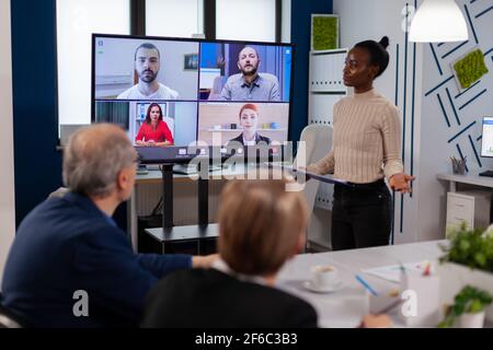 Black manager donna che parla con colleghi in remoto in videochiamata sullo schermo tv, presentando nuovi partner commerciali. Uomini d'affari che parlano con la webcam, fanno la conferenza in linea partecipano al brainstorming del Internet, discussione dell'ufficio di distanza. Foto Stock