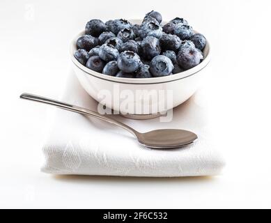 Mirtilli freschi e crudi con gocce d'acqua. In ciotola su tovagliolo bianco con cucchiaio e pronto a mangiare. Isolato con spazio di copia. Foto Stock