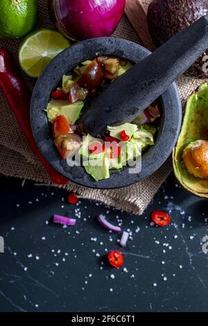 Preparazione di guacamole in un tradizionale mortaio di pietra con tutti i suoi ingredienti (avocado tritato, lime, cipolla, pomodori e peperoncini). Appetizin Foto Stock