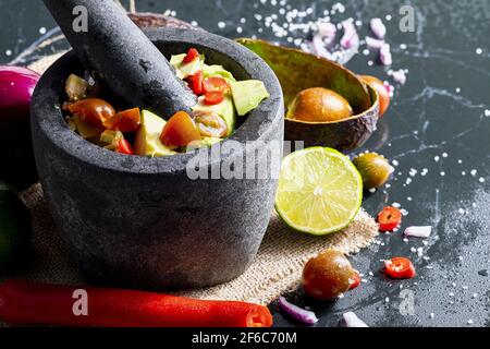 Preparazione di guacamole in un tradizionale mortaio di pietra con tutti i suoi ingredienti (avocado tritato, lime, cipolla, pomodori e peperoncini). Appetizin Foto Stock