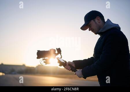 Giovane uomo che riprende con macchina fotografica e gimbal. Videografo in piedi contro la città all'alba. Praga, Repubblica Ceca Foto Stock