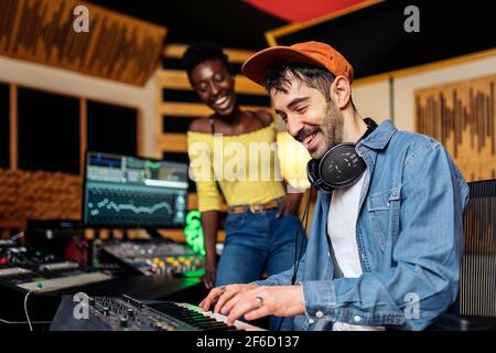 Foto d'inventario di donna nera felice in studio musicale e produttore maschile utilizzando una tastiera elettronica per pianoforte. Foto Stock