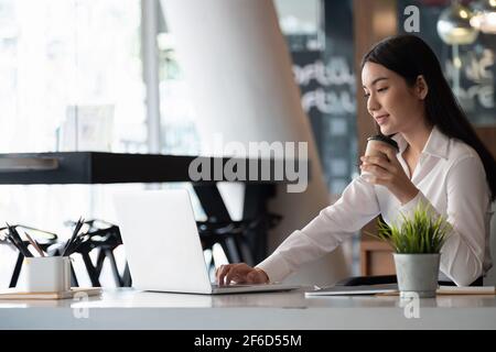 Lavoro remoto. Donna asiatica che lavora a distanza sul suo laptop. Una ragazza brunette in una camicia bianca che fa note durante un briefing online di affari a casa sua Foto Stock