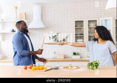 Falso torneo divertente. Ragazza e ragazzo afro-americano misto-razza coppia giocando con un Rolling pin, combattendo nella cucina moderna, mentre la cucina di preparare pizza fatta in casa con verdure Foto Stock