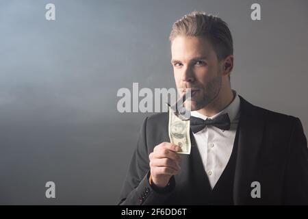 uomo d'affari che illumina il sigaro dalla banconota del dollaro mentre guardando la macchina fotografica su sfondo grigio con fumo Foto Stock