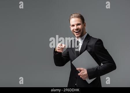 ridendo uomo d'affari che punta con le dita mentre tiene il laptop isolato nero Foto Stock