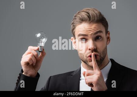 un uomo d'affari entusiasta che tiene una lampadina mentre mostra un gesto segreto isolato su grigio Foto Stock