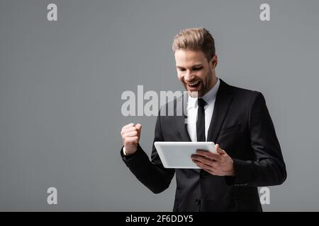 uomo d'affari entusiasta che mostra il gesto di vincere tenendo il tablet digitale isolato su grigio Foto Stock