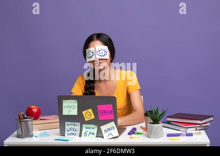 Donna stanca seduta alla scrivania con note appiccicose sugli occhi Foto Stock