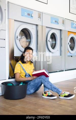 Giovane donna latino-americana in attesa che i suoi vestiti siano lavati in lavanderia self-service. Lei sorride e ha un libro nelle sue mani. Foto Stock