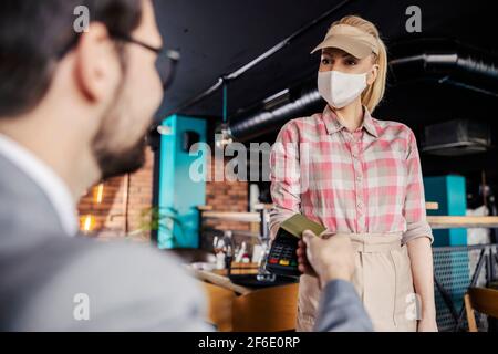 Pagamento dell'ordine online durante la corona. Un uomo si siede in un ristorante e paga il conto con una carta al terminale tenuto da una bella cameriera in un pa Foto Stock