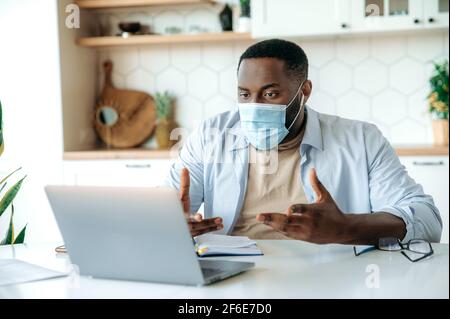 Lavoro da casa durante la quarantena. Freelance o manager africano americano maschile, indossando una maschera medica protettiva, lavorando in remoto, chattando con colleghi o clienti tramite videoconferenza con un computer portatile Foto Stock