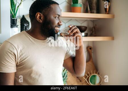 Afroamericano attraente maschio che beve un'acqua pura, seguire uno stile di vita sano. Bel ragazzo si sente sete, bere ogni giorno quantità di liquido puro, in piedi in cucina, concetto di salute Foto Stock