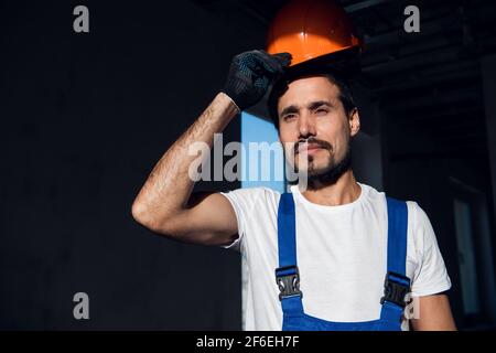 Un riparatore regola un elmetto arancione sulla testa. Indossa una tuta da lavoro Foto Stock