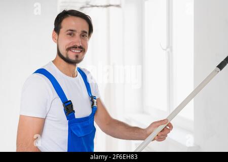 Colpo medio, handyman in abiti da lavoro blu dipinge parete in appartamento Foto Stock