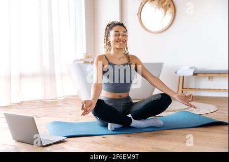 Calma cute giovane donna afroamericana in sportswear fa yoga e meditazione seduta nella posizione del loto, a casa su un tappetino fitness, conduce uno stile di vita sano, si prende cura della sua salute, sorrisi Foto Stock