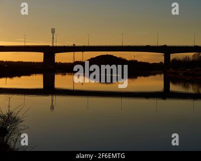 Un ponte scolpito attraverso un tranquillo fiume al tramonto, con l'intricata ironia delle luci di strada e dei riflettori catturati contro il cielo dorato. Foto Stock