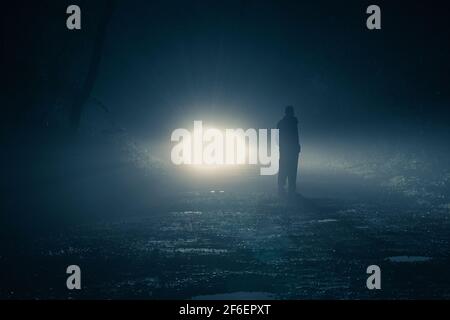 I fari dell'automobile illuminano indietro un uomo e gli alberi su una notte fogy spooky in una foresta. Foto Stock