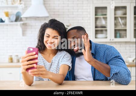 Felice coppia afroamericana che usa lo smartphone per videochiamare la famiglia o gli amici seduti in cucina a casa. Giovani sposi multirazziali salutano i seguaci, registrando video blog Foto Stock