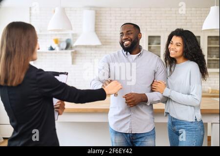Concetto di raggiungimento degli obiettivi. Felice coppia afroamericana che ottiene le chiavi dalla propria proprietà dall'agente immobiliare. Gli sposi multirazziali si trasferiscono, spostandosi in un nuovo appartamento Foto Stock