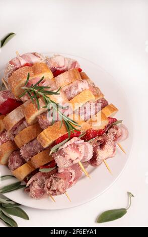 Spiedini crudi rustici, pronti a cucinare, con tacchino, salsiccia, maiale, verdure, erbe e fette di pane su sfondo bianco. Primo piano. Foto Stock