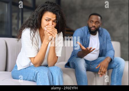 Una donna biraciale frustrata piangente si siede in primo piano, depressa e infelice, un uomo afro-americano fuori fuoco sullo sfondo. La coppia multirazziale è vicina al divorzio. Concetto di abuso domestico Foto Stock