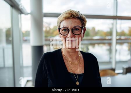 Primo piano di una donna aziendale senior in ufficio che guarda la fotocamera e sorride. Donna matura che indossa occhiali in ufficio. Foto Stock