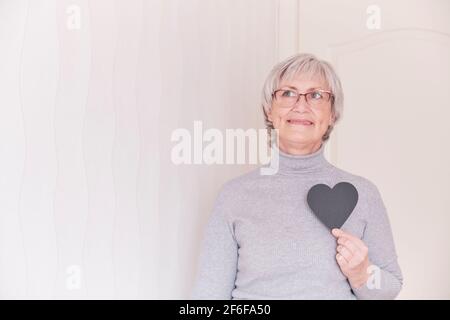 Un ritratto di una donna caucasica anziana con i capelli grigi corti che portano gli occhiali tenendo in mano un cuore nero. Foto Stock